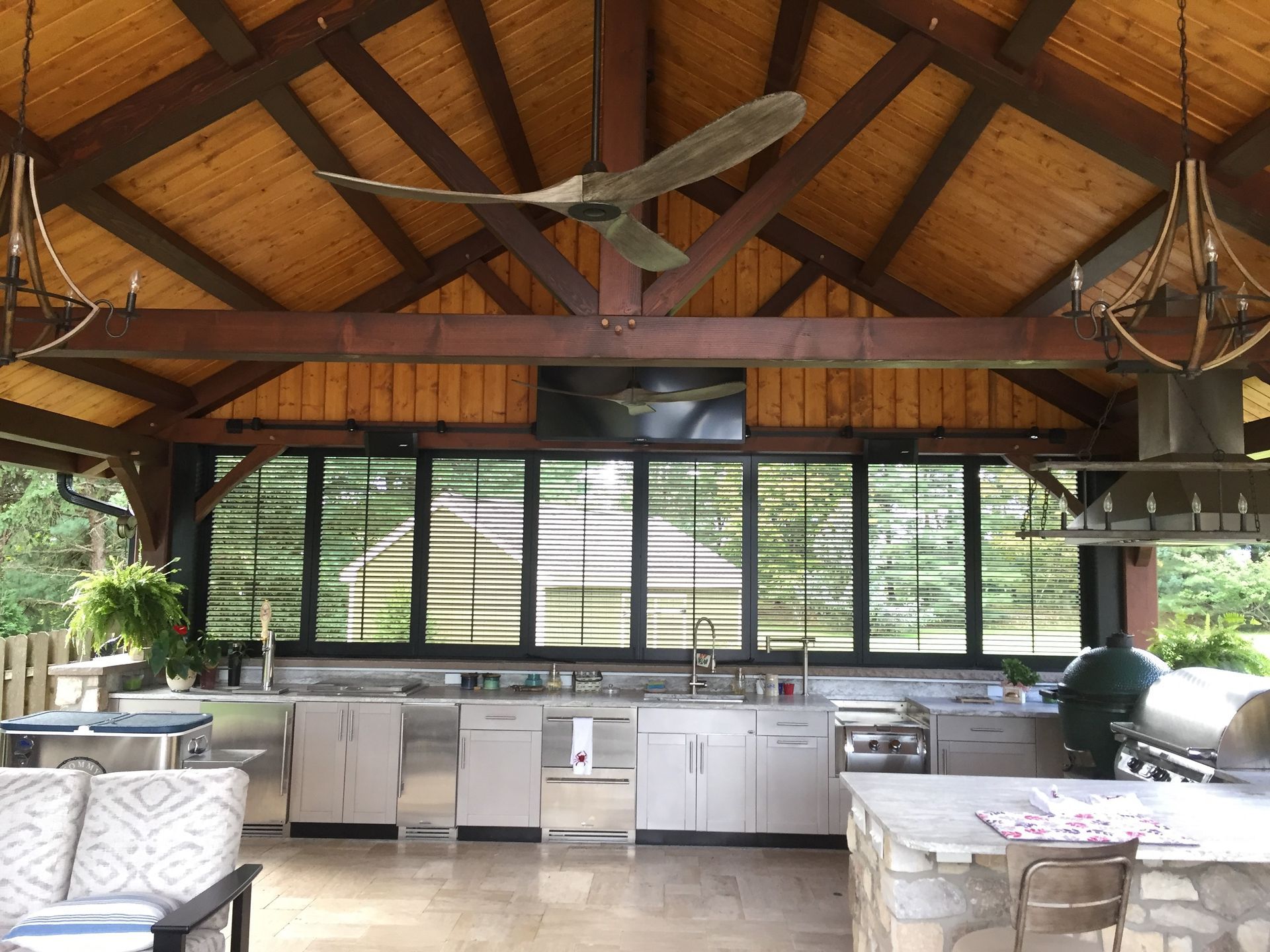 Open-air kitchen with wooden vaulted ceiling, large windows, stainless counters, and seating overlooking greenery