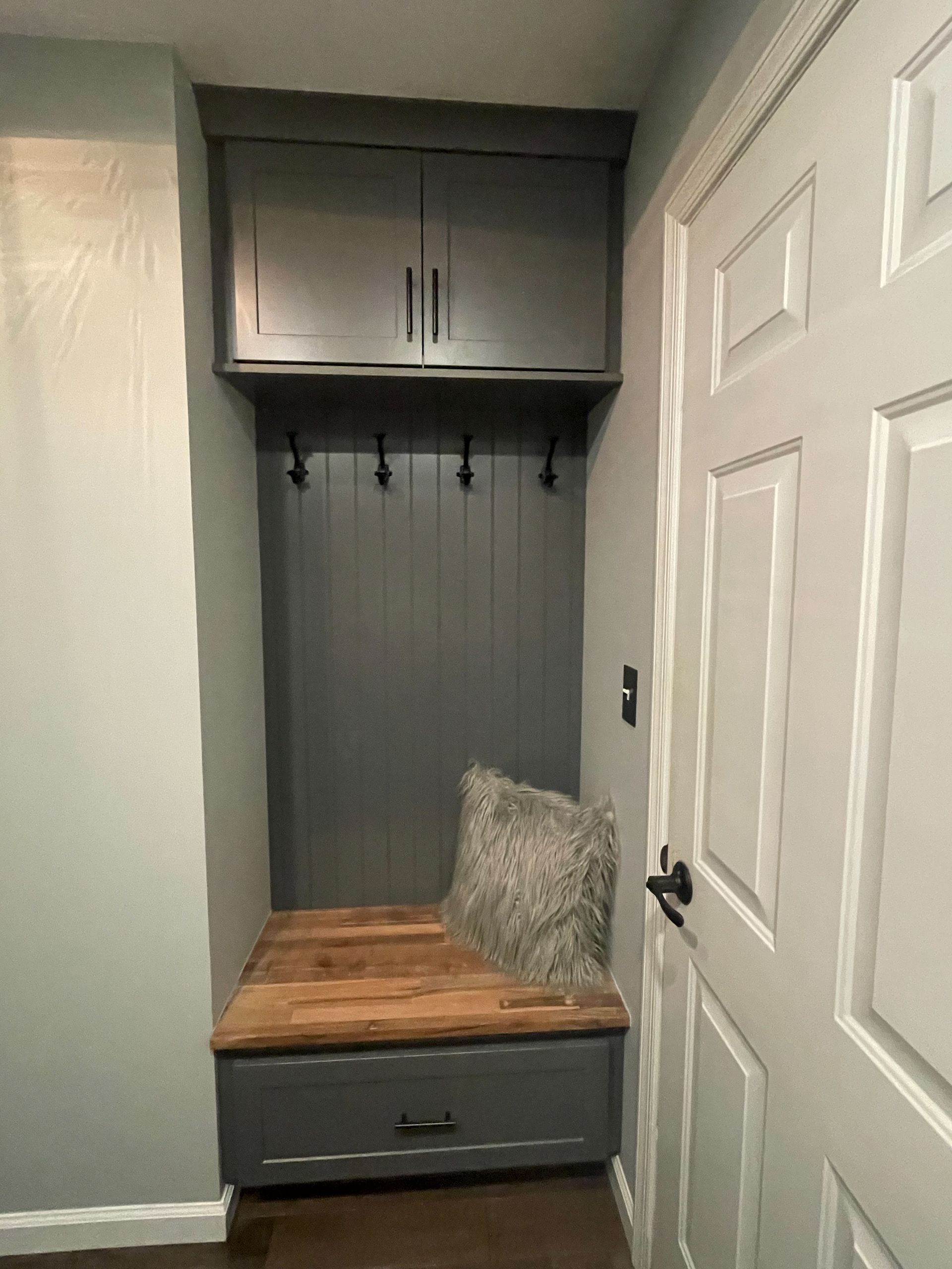 Small entryway bench with wood seat, gray cushion, hooks, and overhead cabinet in a narrow alcove.
