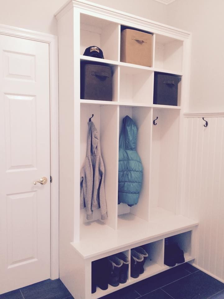White entryway storage bench with cubbies holding coats, hats, boxes, and shoes beside a closed door.