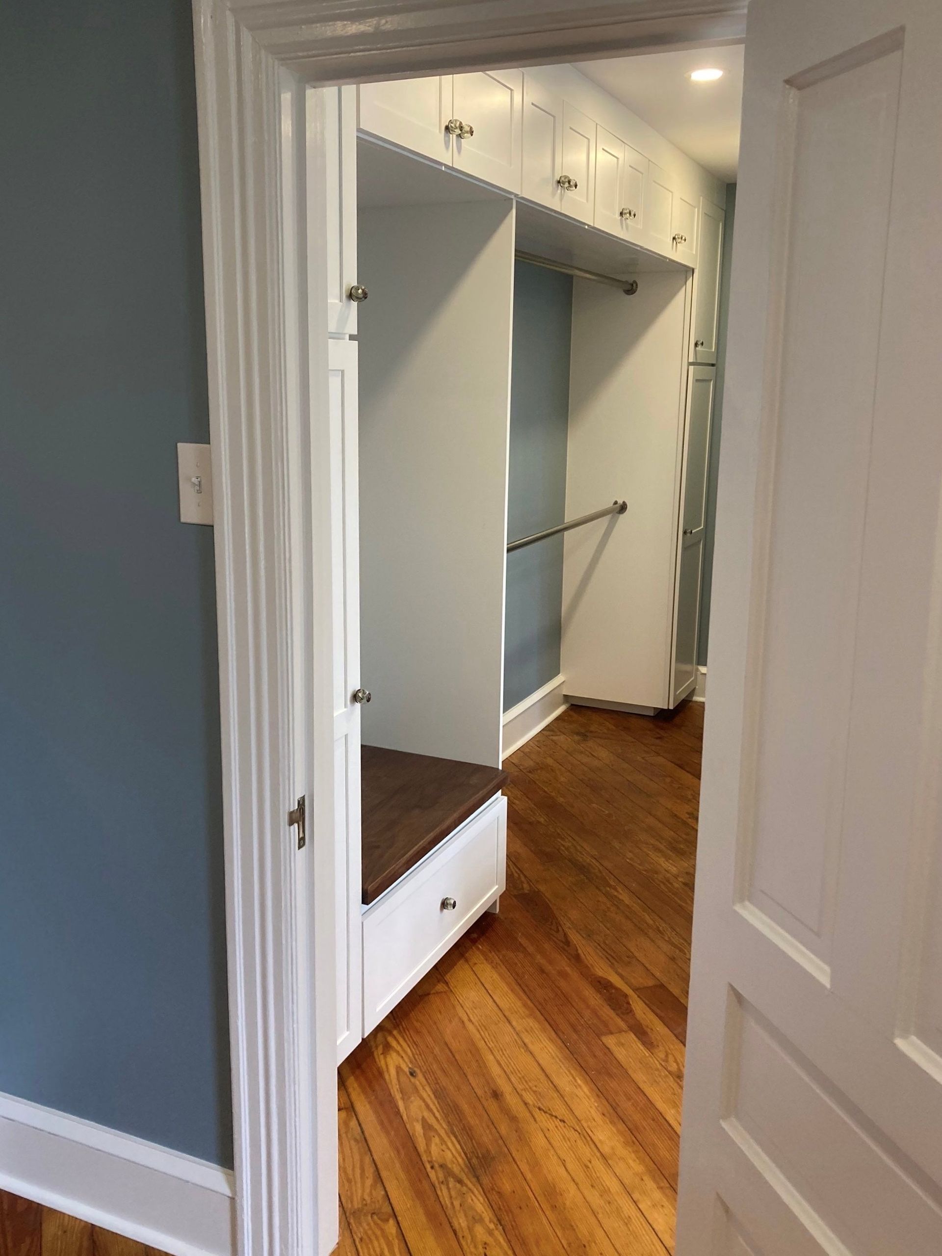 Open walk-in closet with white built-ins, hanging rod, drawer, and wood floor, viewed through a doorway