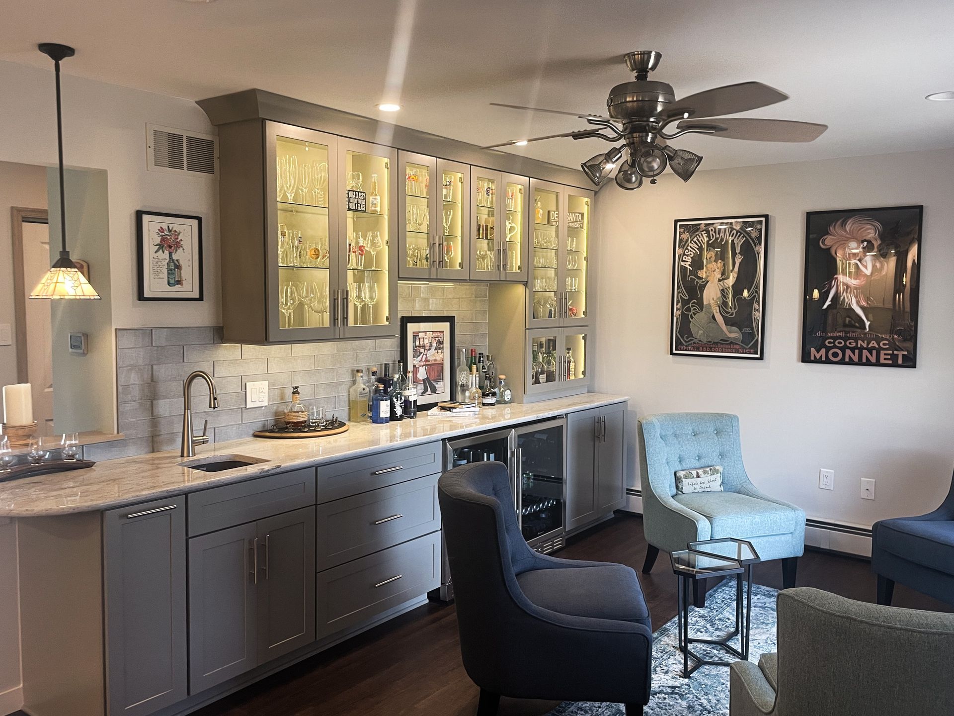 Modern kitchen and living area with dark cabinets, lit glass cupboards, and two chairs by a wall-mounted TV.