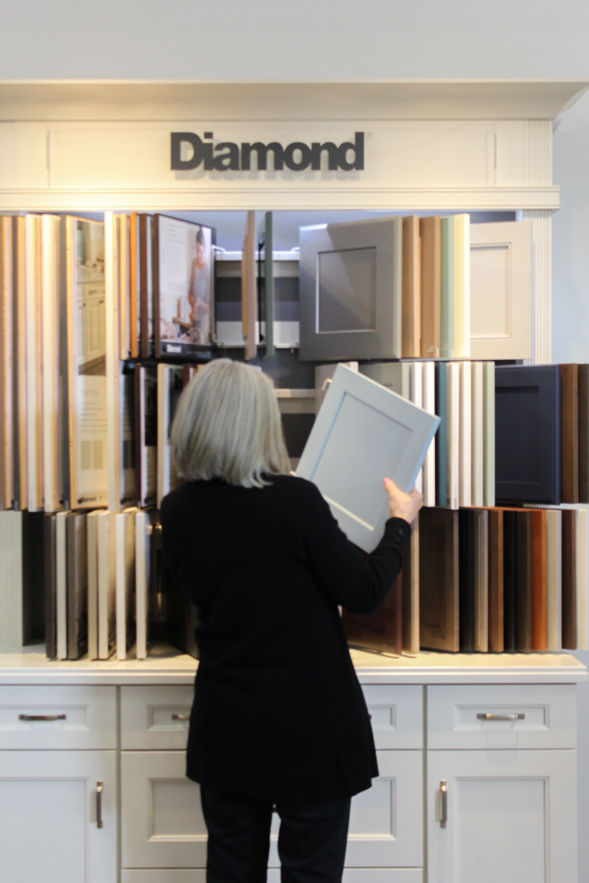Person holding a white cabinet door in a showroom labeled “Diamond,” with cabinetry samples on display