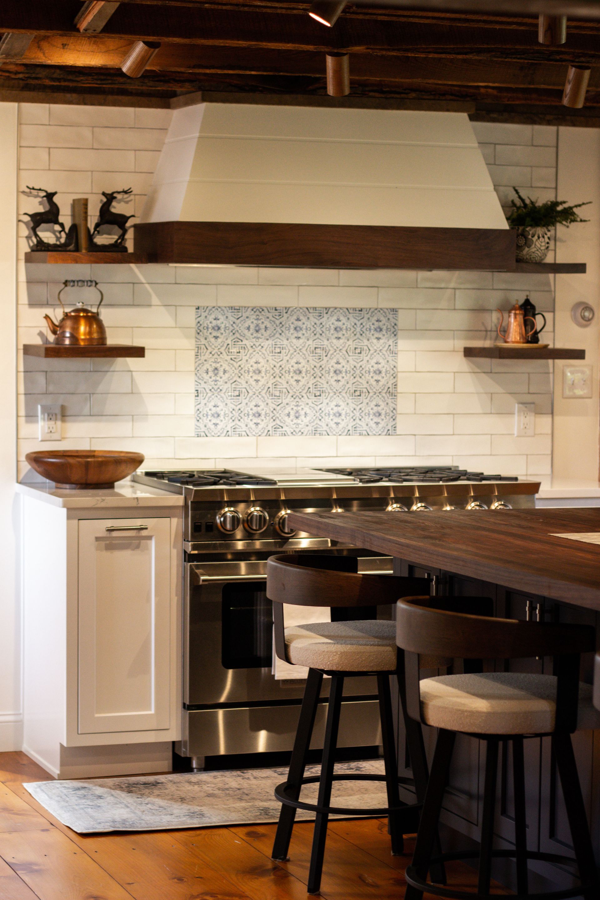 Cozy kitchen with stove, wooden island, open shelves, and decorative backsplash beneath a wood beam.