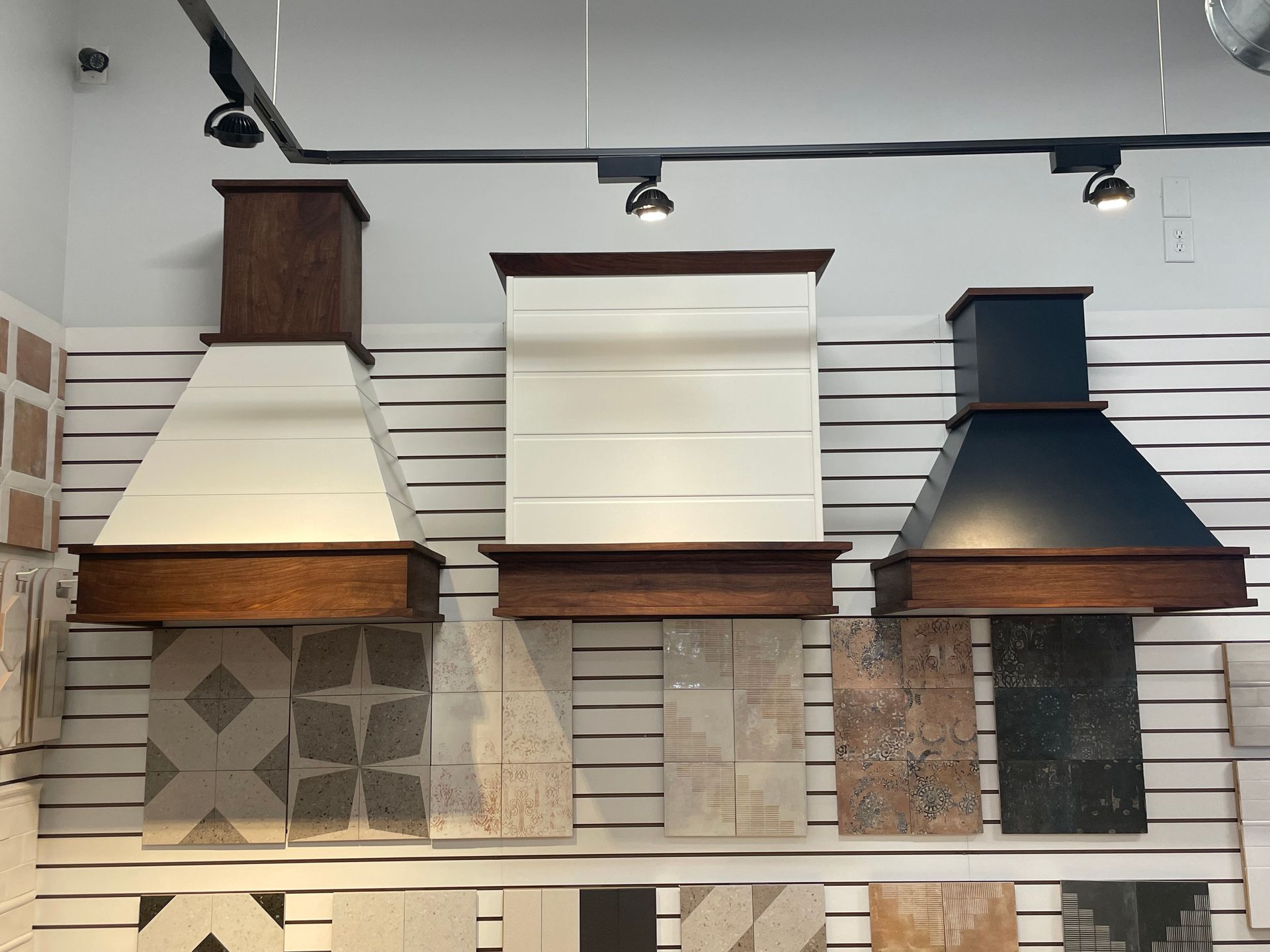 Three wall-mounted range hoods in a showroom, displayed in dark wood, white, and navy finishes.