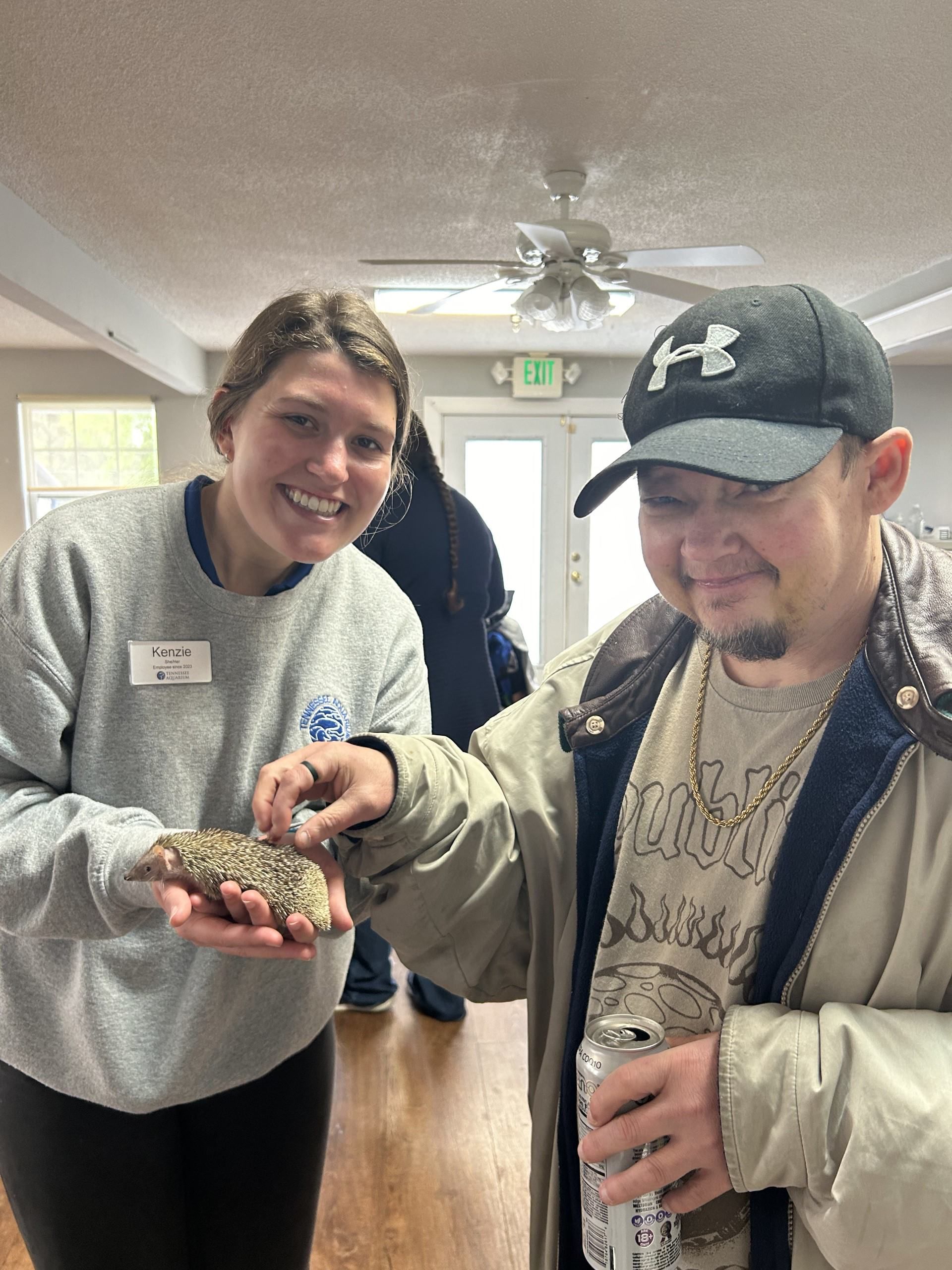 Two people smile while holding a small, spiky animal in a bright, indoor community space with a ceiling fan.