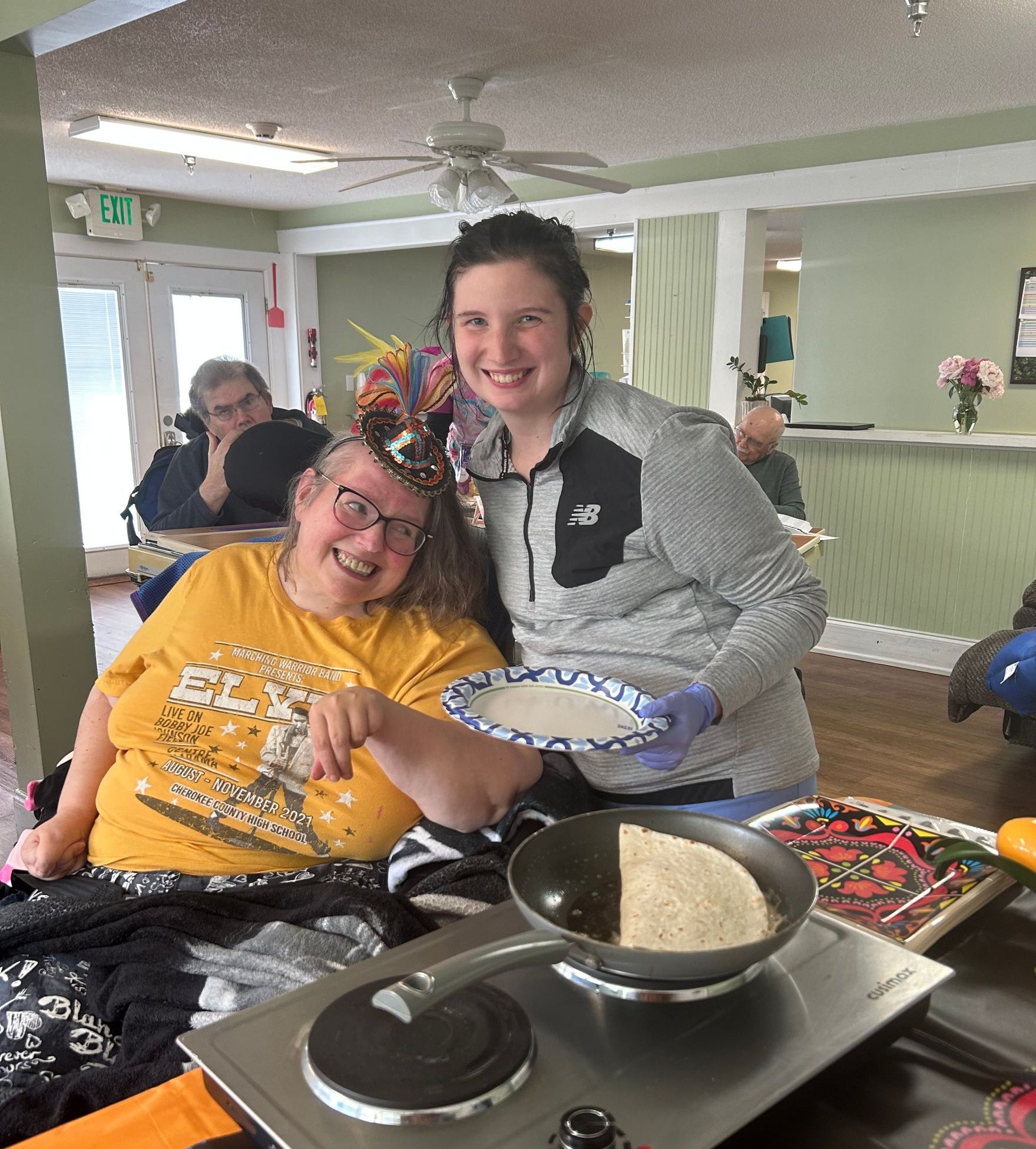 Two women cooking pancakes, one in a wheelchair, smiling. One holds a pancake, the other points to the pan on the hot plate.