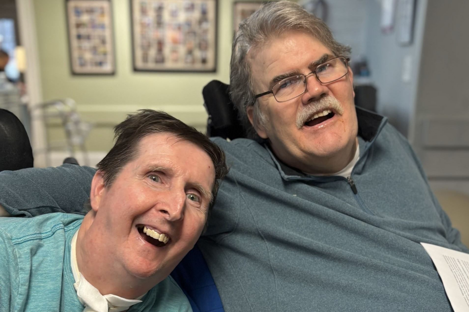 Two people in wheelchairs smiling together in a room with framed pictures on the wall.