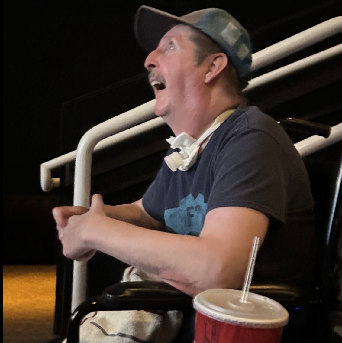 Man in a wheelchair, mouth open, looking up with a surprised expression. He's in a theater with a drink.