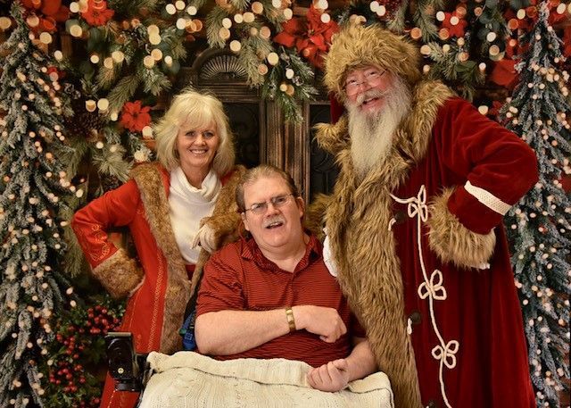 Man in wheelchair with Santa and helper in front of a Christmas backdrop.