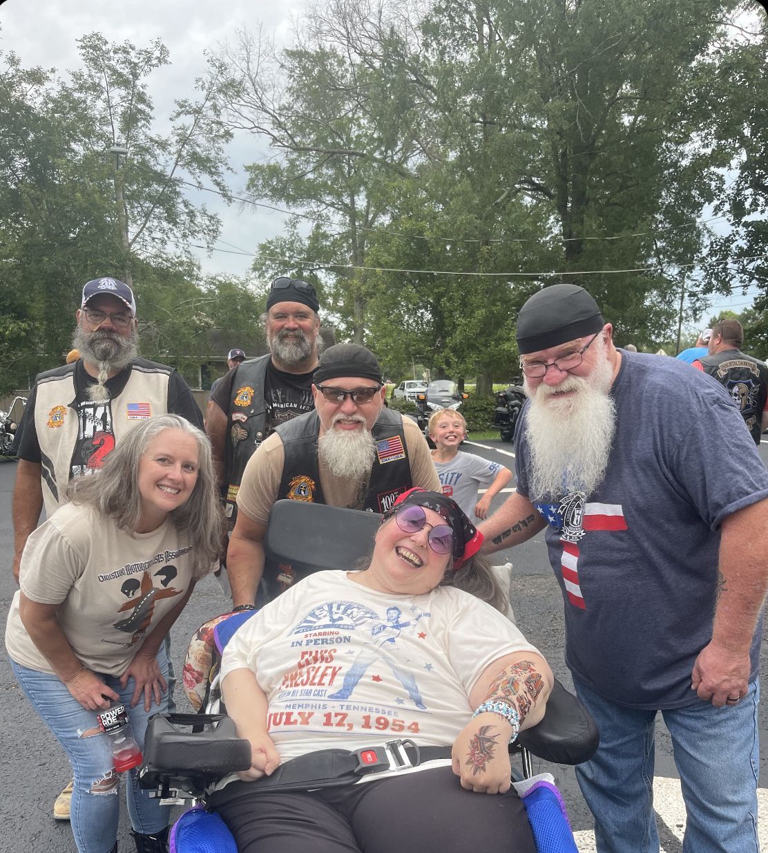 Group of people, some with beards, pose around a woman in a wheelchair. Outdoors, near a tree line.