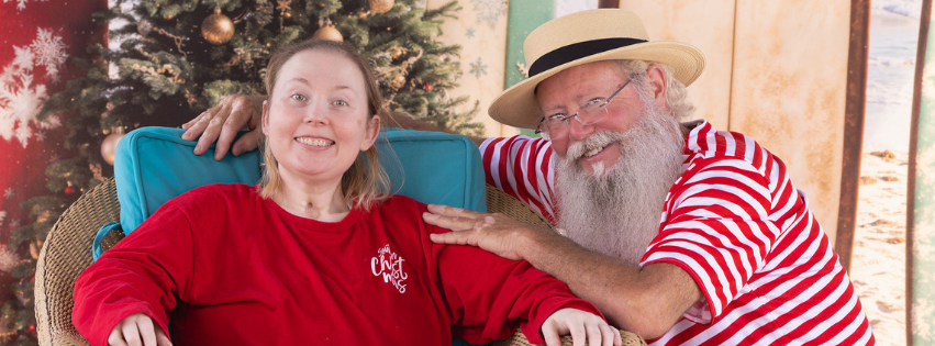A woman in a red shirt smiles next to Santa, who wears a striped shirt, hat, and has a beard.