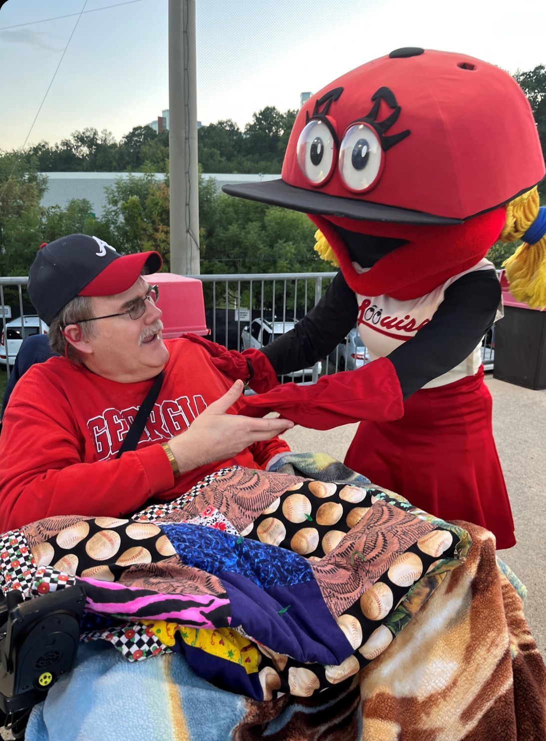 Man in a wheelchair shakes hands with a red softball mascot wearing a cap. Outdoor setting.