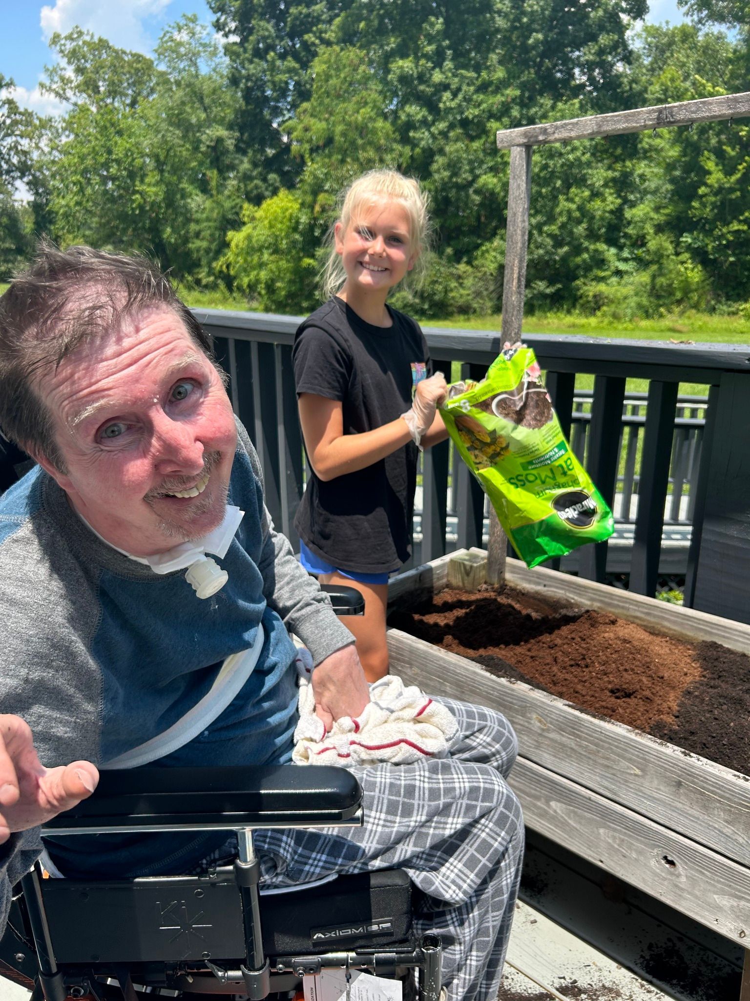 A man in a wheelchair smiles as a girl pours soil into a raised garden bed on a deck. Sunny day.