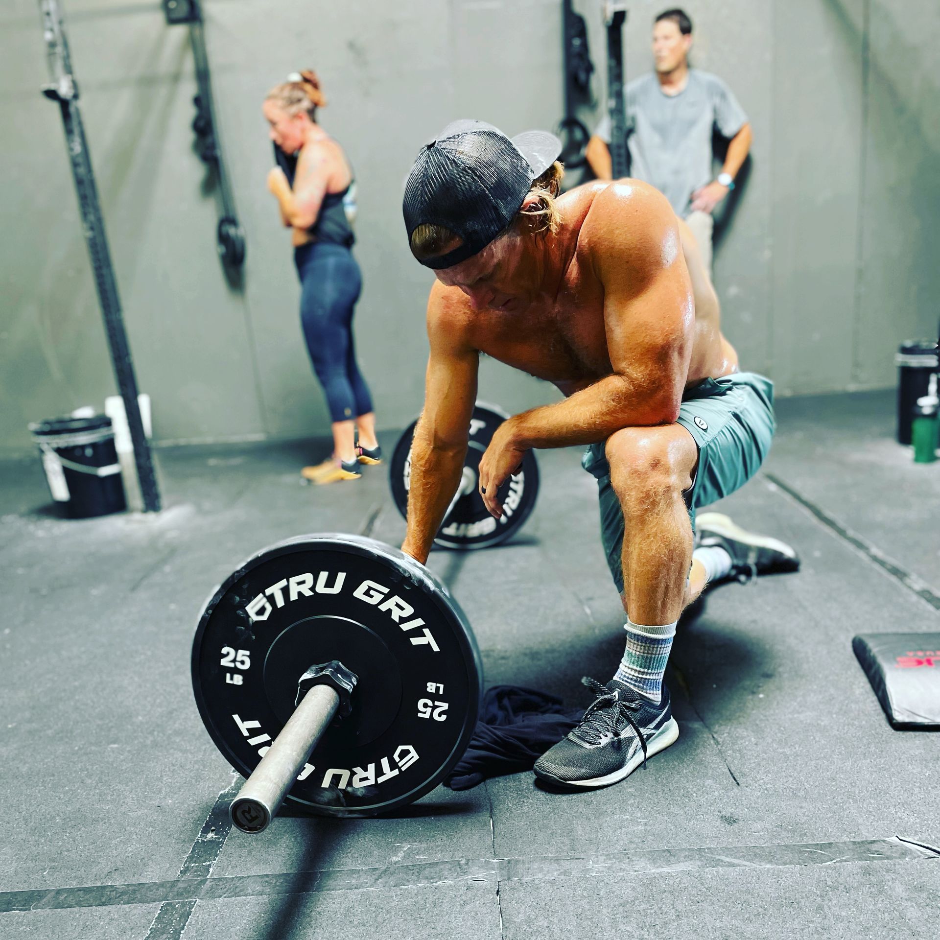 Athlete resting on a gym floor beside a loaded barbell, with two people visible in the background.