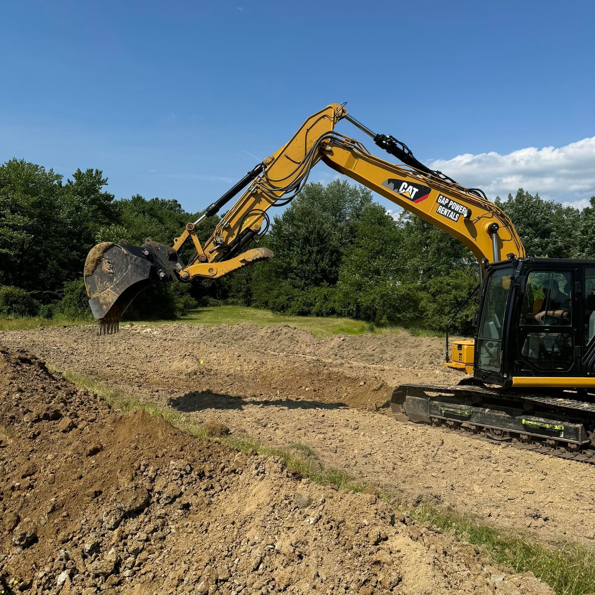 A yellow Caterpillar excavator sits on a dirt construction site against a backdrop of trees and a blue sky.