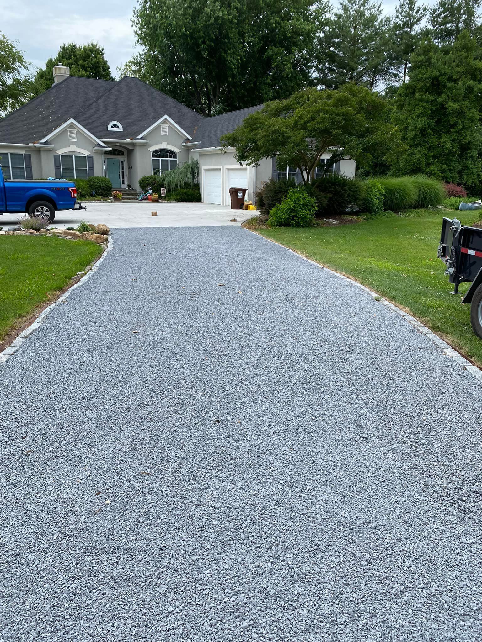 A light-colored, gravel driveway leads toward a gray suburban house with a two-car garage and a blue truck parked outside.