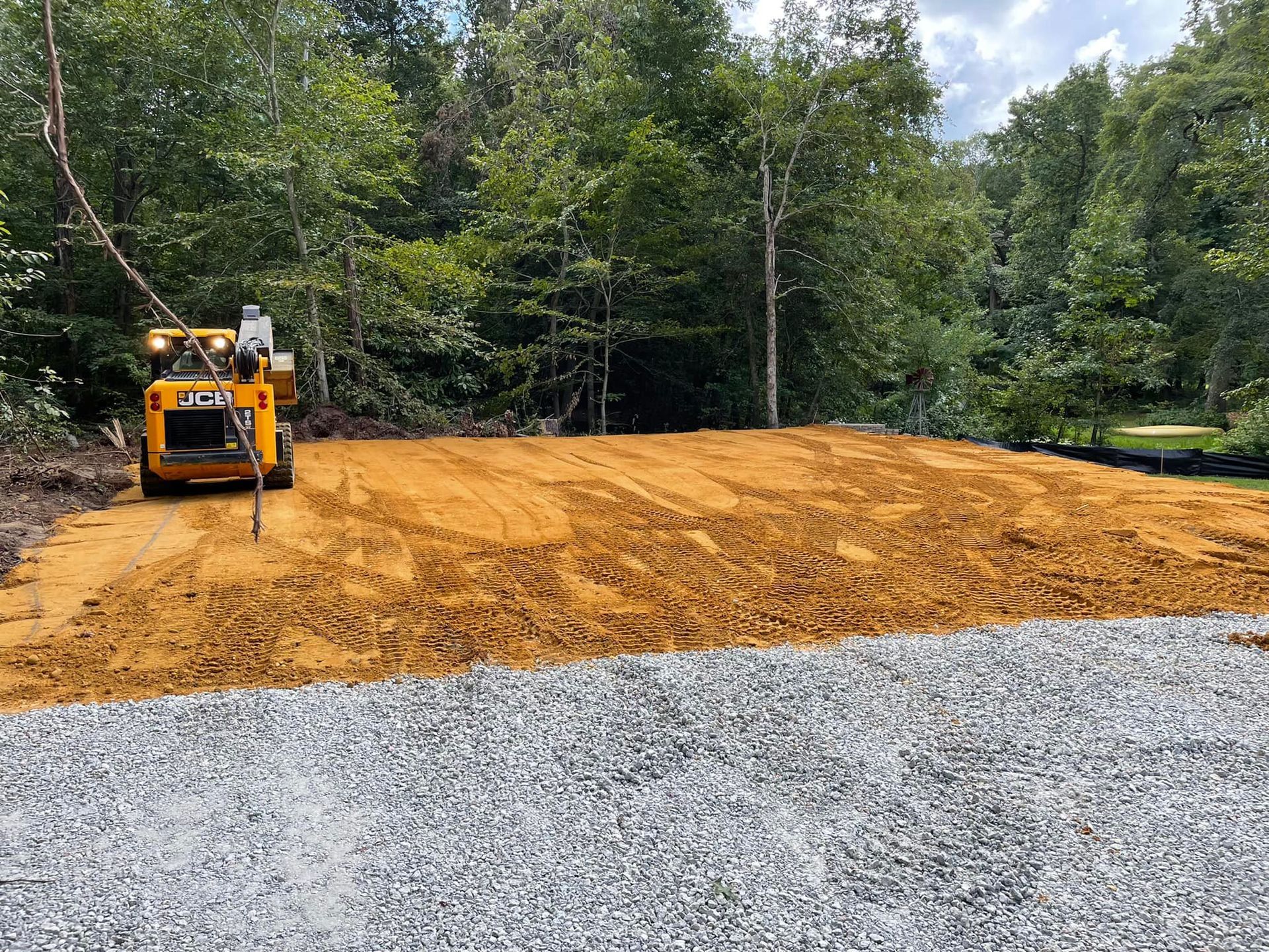 A yellow skid steer tractor spreads dirt on a site cleared of trees, with a gravel foreground.