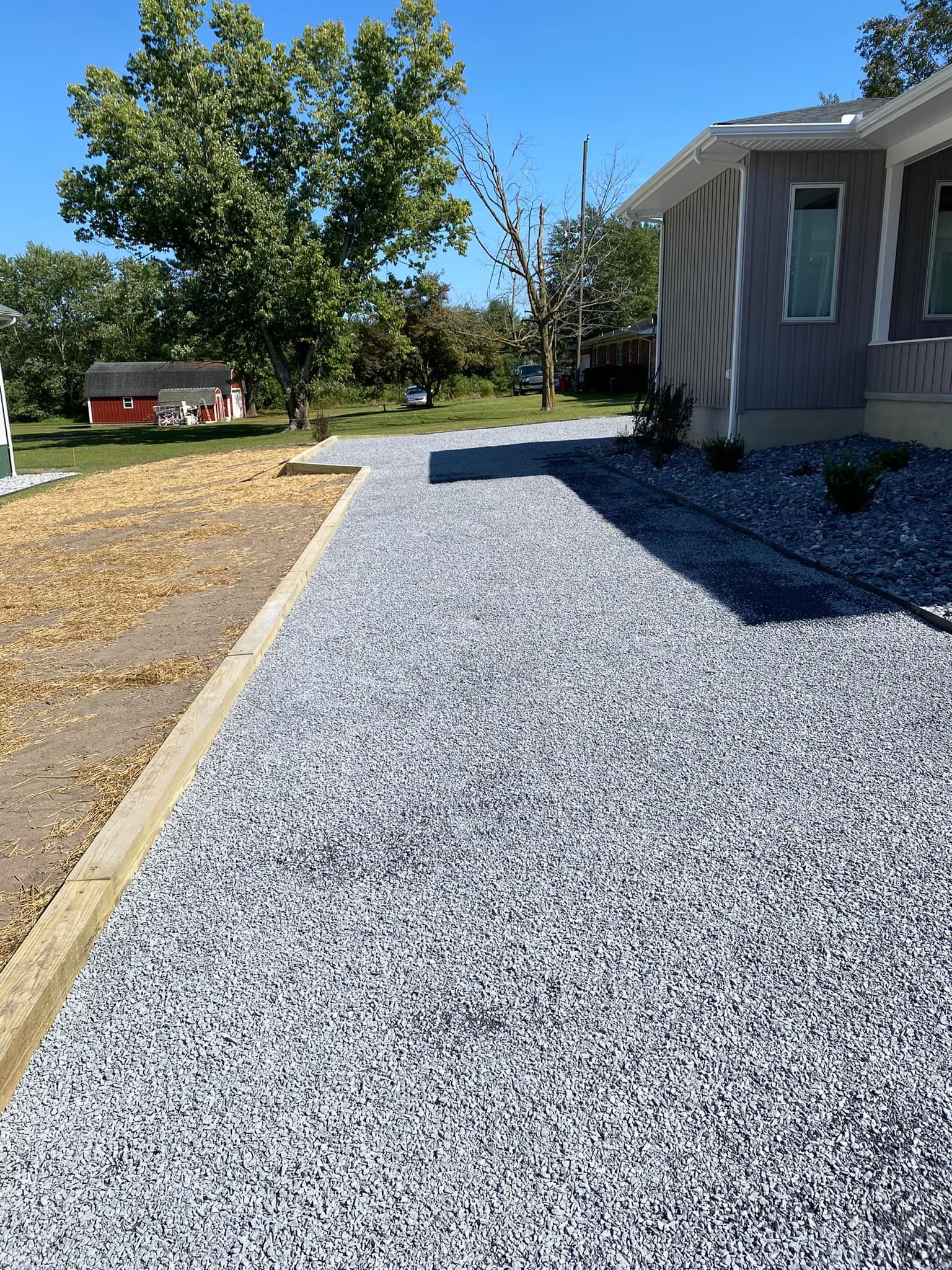A gray stone driveway leads toward a house with gray siding under a clear blue sky, next to a yard with a red shed.