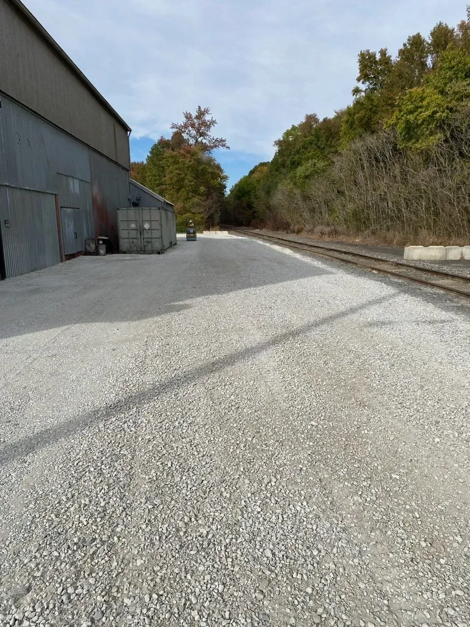 A gravel lot beside a metal warehouse building, bordered by a railroad track and a line of trees under a cloudy sky.