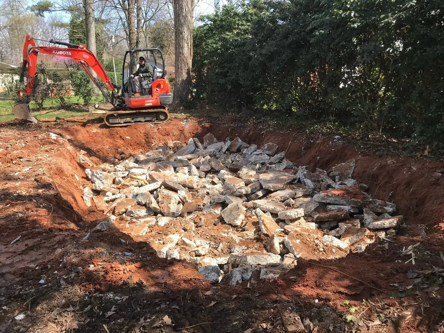 An orange excavator parks next to a large, rectangular pit filled with broken concrete rubble in a wooded area.