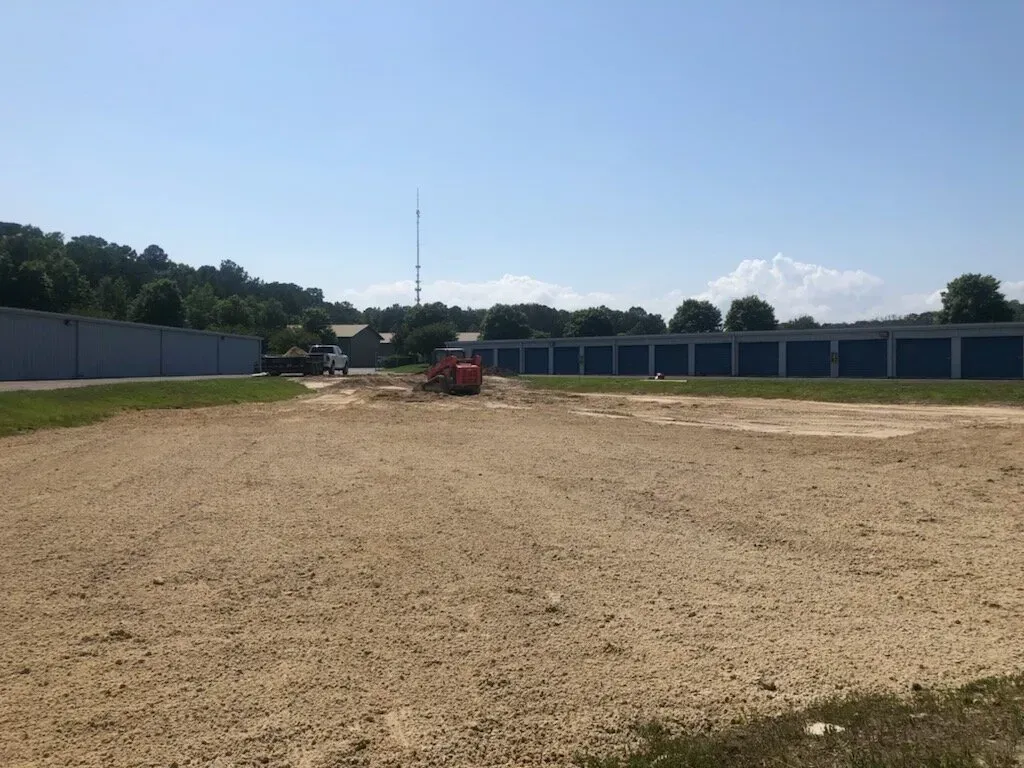Construction equipment clears dirt on a sandy lot between two rows of blue storage units under a clear blue sky.