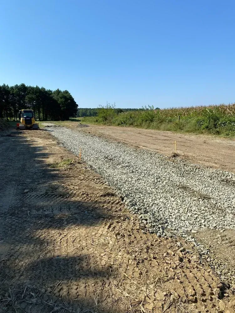 A construction vehicle sits on a gravel path bordering a field under a clear blue sky.