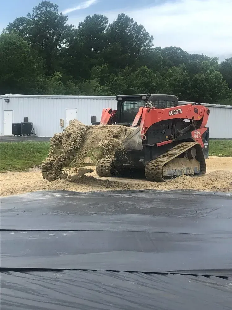 An orange tracked skid steer loader moves a scoop of dirt across a construction site with a black ground tarp nearby.