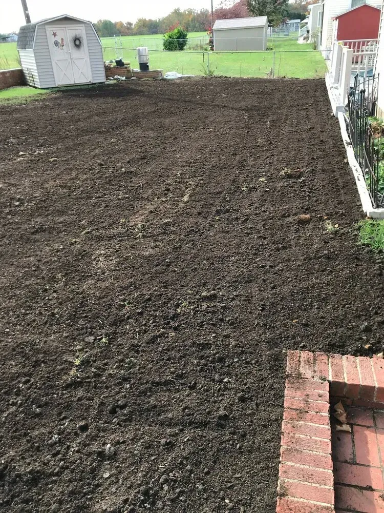 A view of a freshly tilled, dark soil backyard with storage sheds in the background under a cloudy sky.