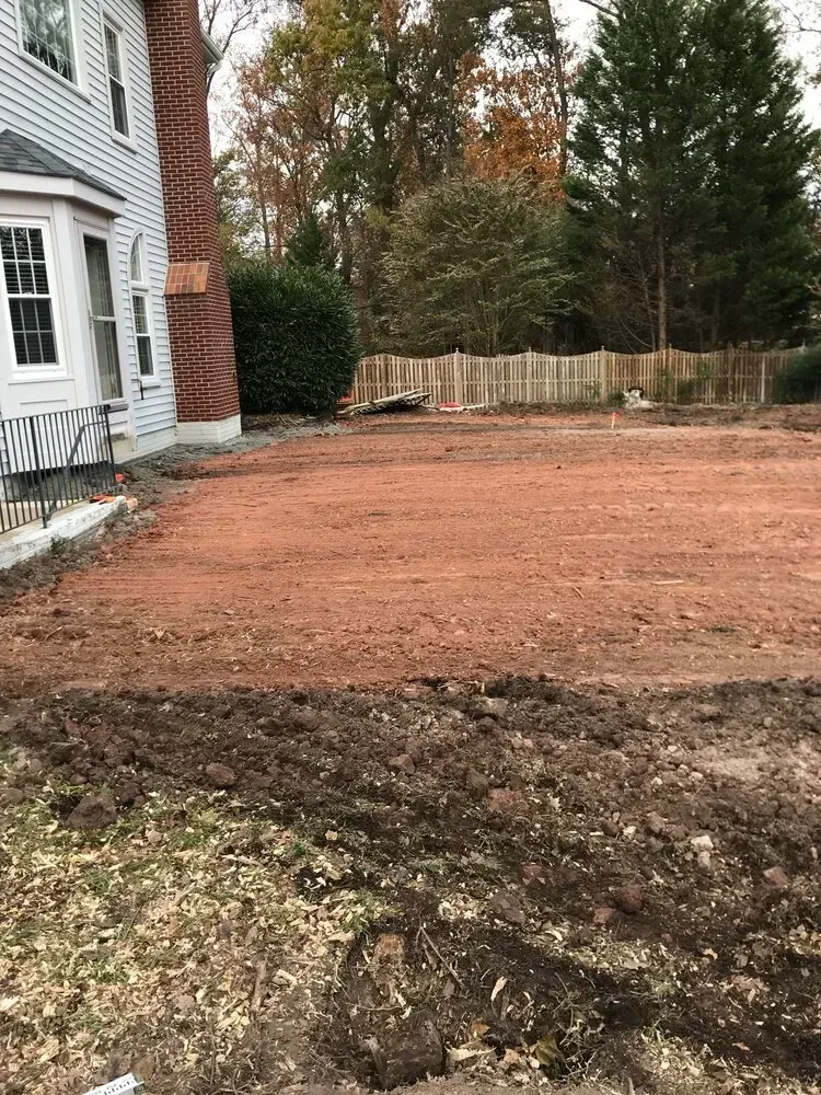 A residential backyard under construction with a large area of leveled red dirt alongside a white house and brick chimney.