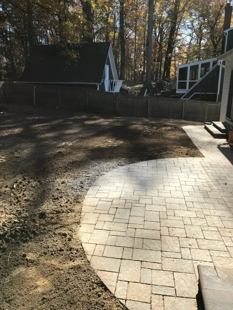 A newly installed light-colored stone patio curves into an unfinished dirt yard with trees and houses in the background.