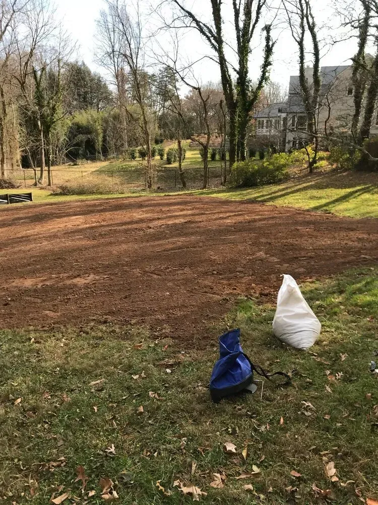 A patch of tilled, bare soil in a grassy yard, with a white bag and blue tarp sitting on the lawn.