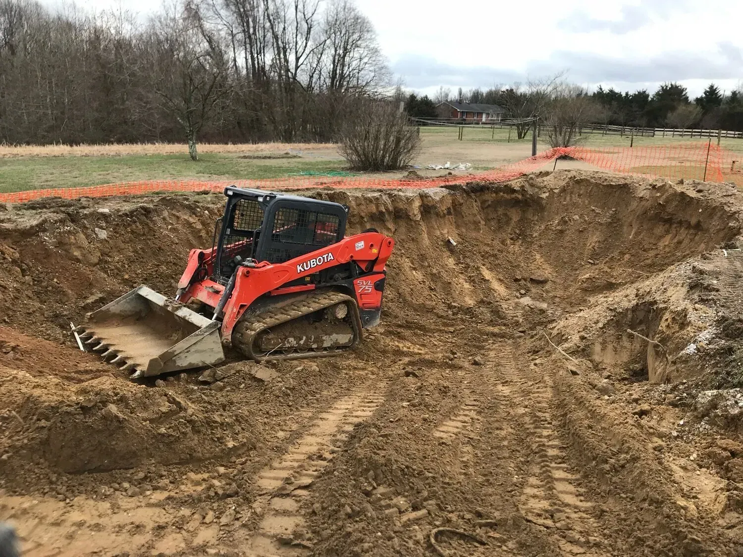 A bright orange skid-steer loader sits inside a large, deep excavation pit on a dirt construction site.