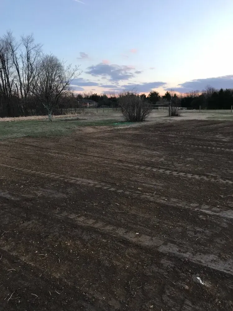 A freshly tilled, dark soil field with tire tracks under a twilight sky with trees in the background.