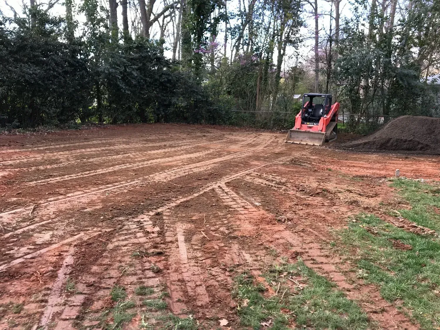 A red skid steer loader levels a red clay construction site with a pile of dirt nearby in a wooded area.