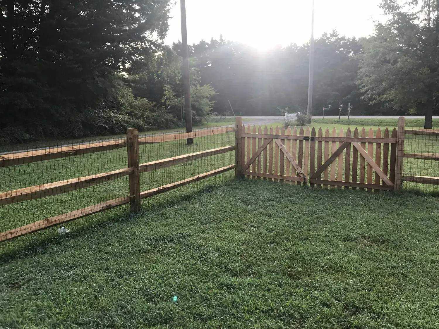 A wooden split-rail fence surrounds a grassy yard, featuring a gated entrance against a background of trees and sky.