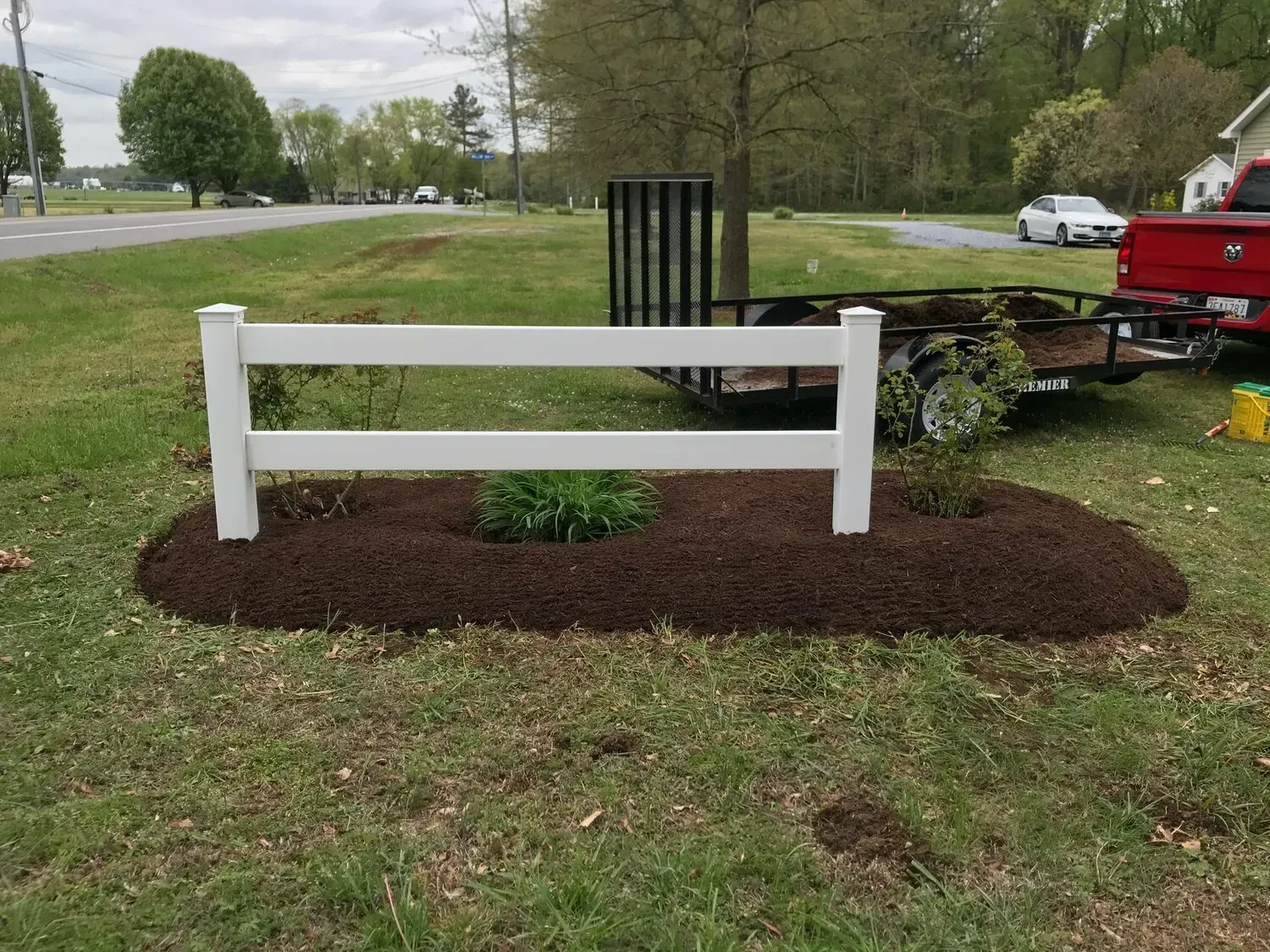A small white two-rail fence sits in a dark mulch bed surrounded by green grass with a utility trailer parked nearby.