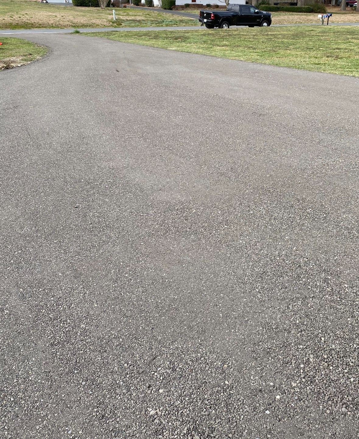 A long, gray gravel driveway curves toward a black pickup truck parked in a grassy residential area.