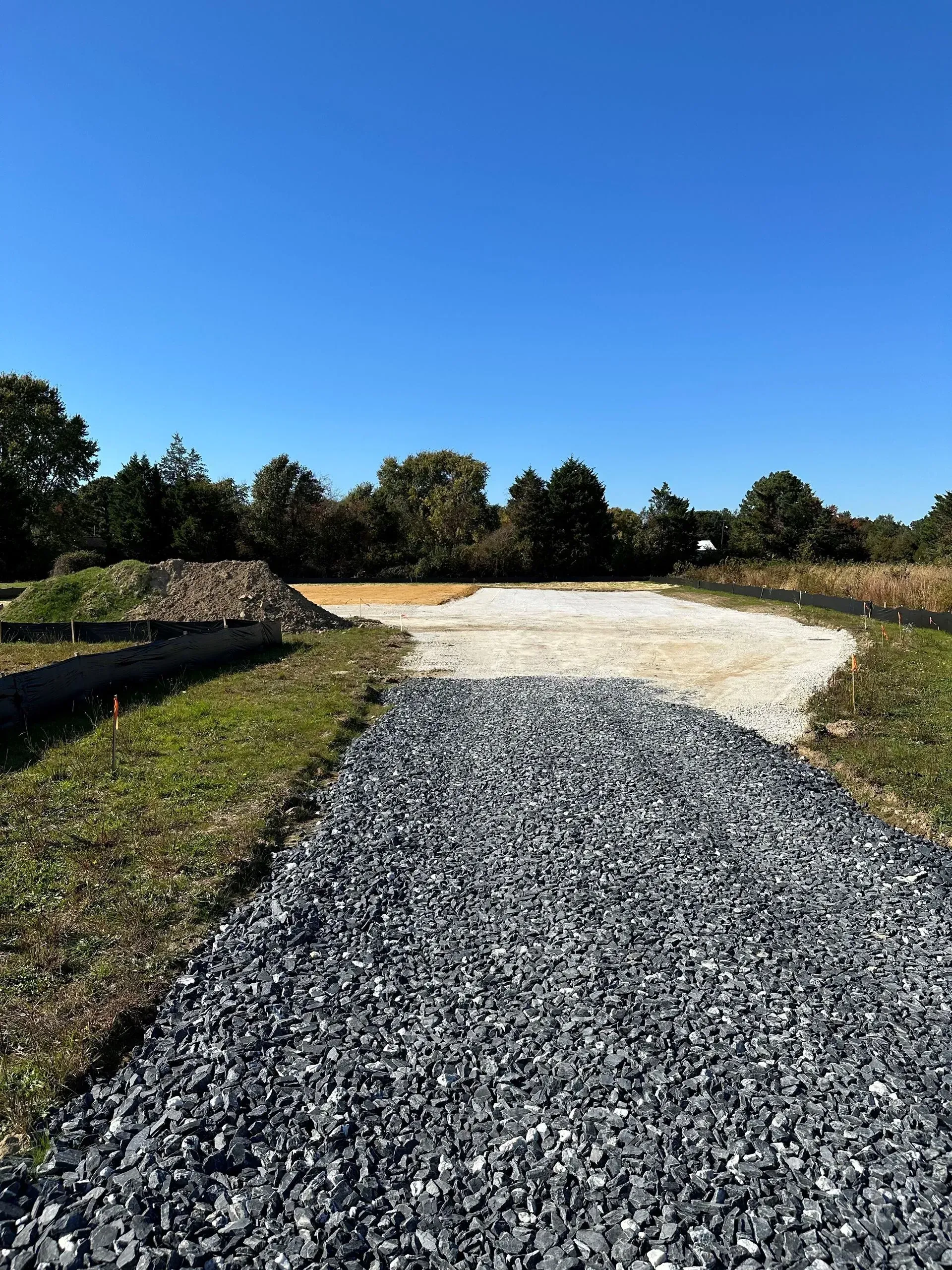 A path transitioning from dark grey gravel to light-colored stone, leading toward trees under a clear blue sky.