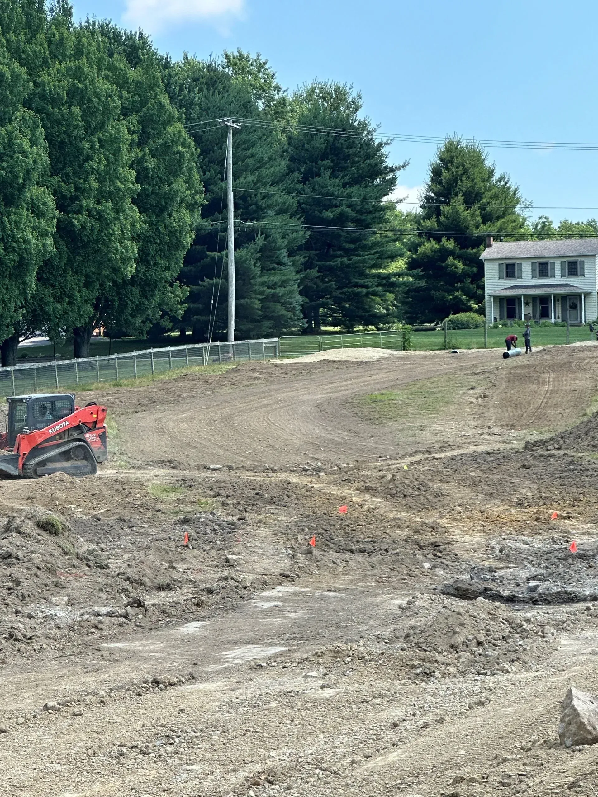 A red skid-steer loader works on a cleared, uneven dirt construction site with trees and a house in the background.