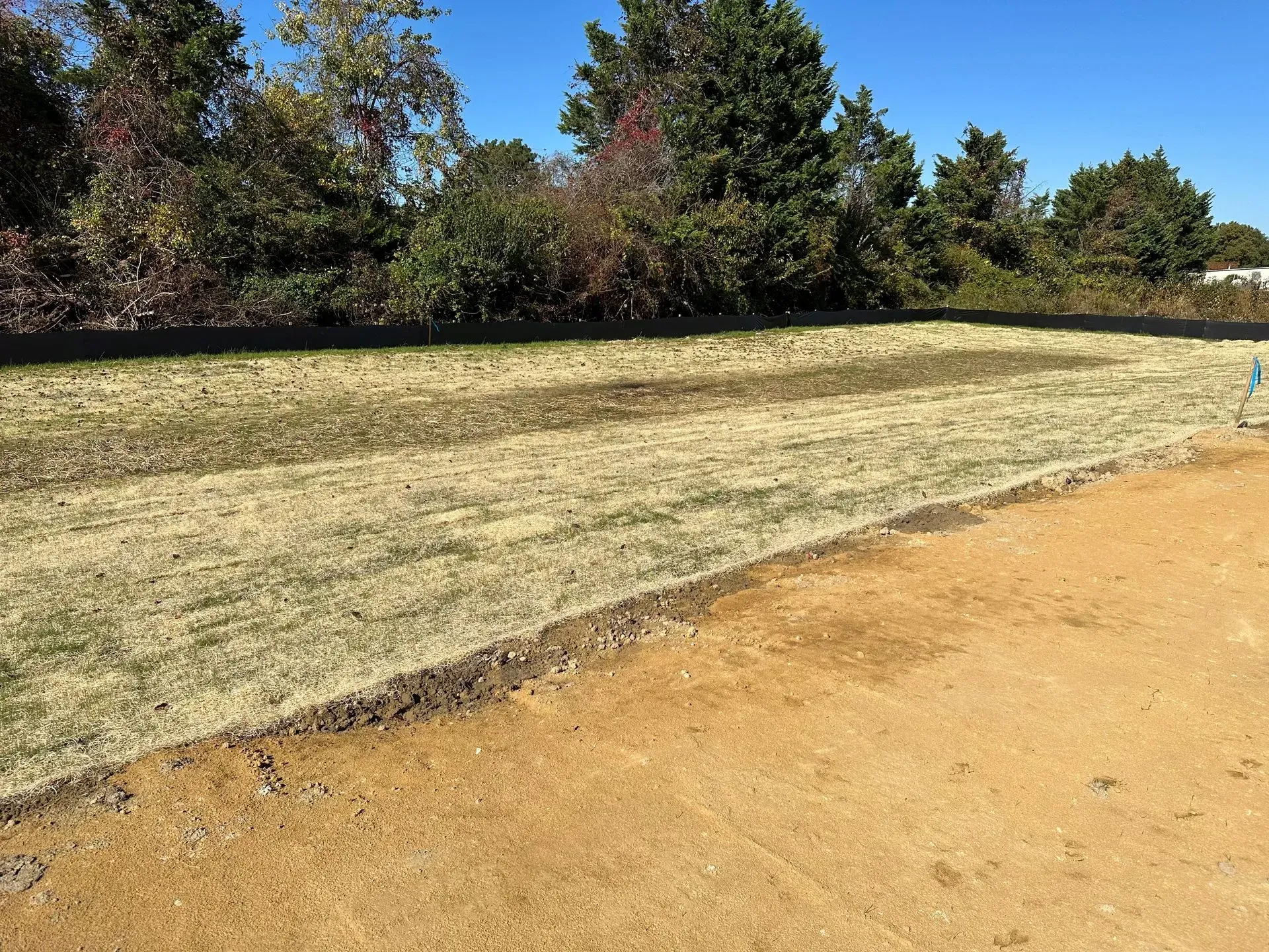 A diagonal view shows a patch of dry, yellowish grass next to a freshly graded area of bare, light-brown soil.