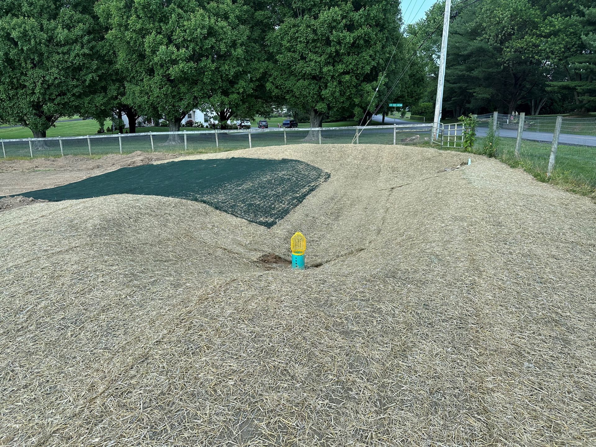 A small circular area of wood chips surrounds a central yellow utility marker in a grassy field with a perimeter fence.