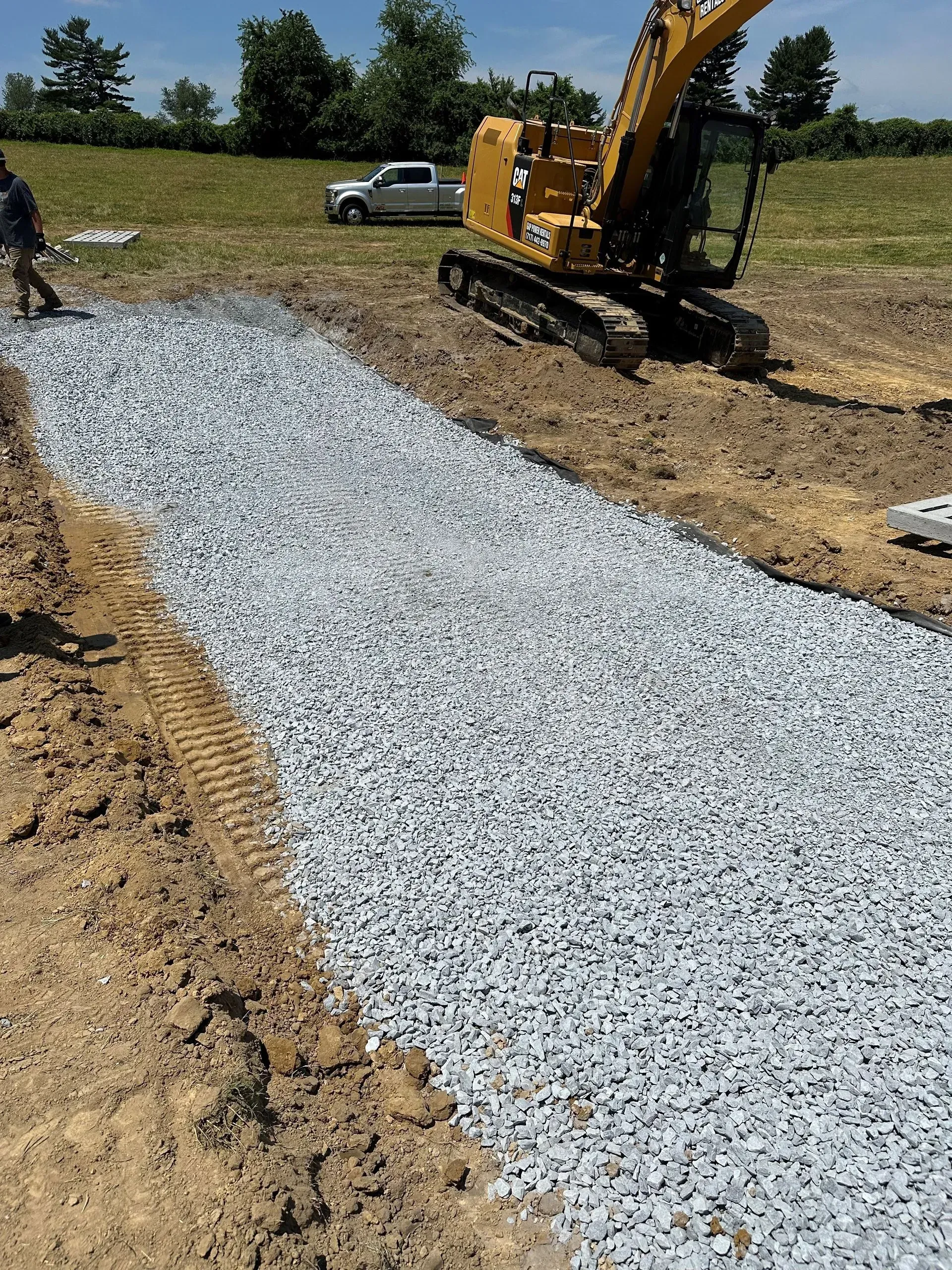 A construction excavator sits near a long, straight trench filled with grey gravel in an open field.