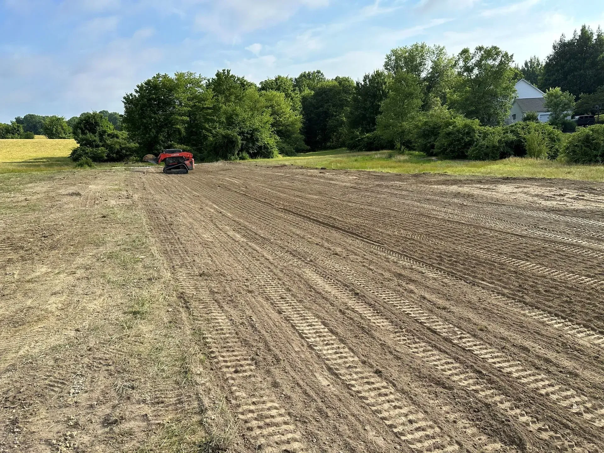 A red tractor tilling a large, dirt-covered field bordered by trees under a bright blue sky.