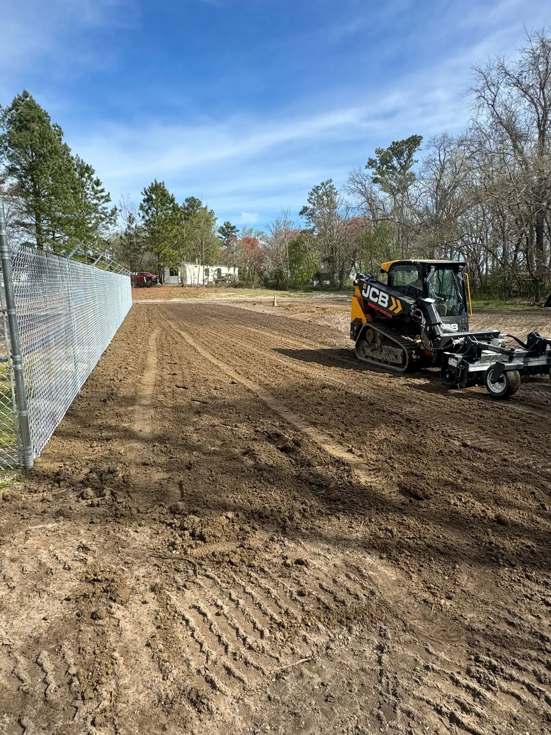 A yellow and black JCB skid steer levels tilled soil next to a chain-link fence under a blue, partly cloudy sky.