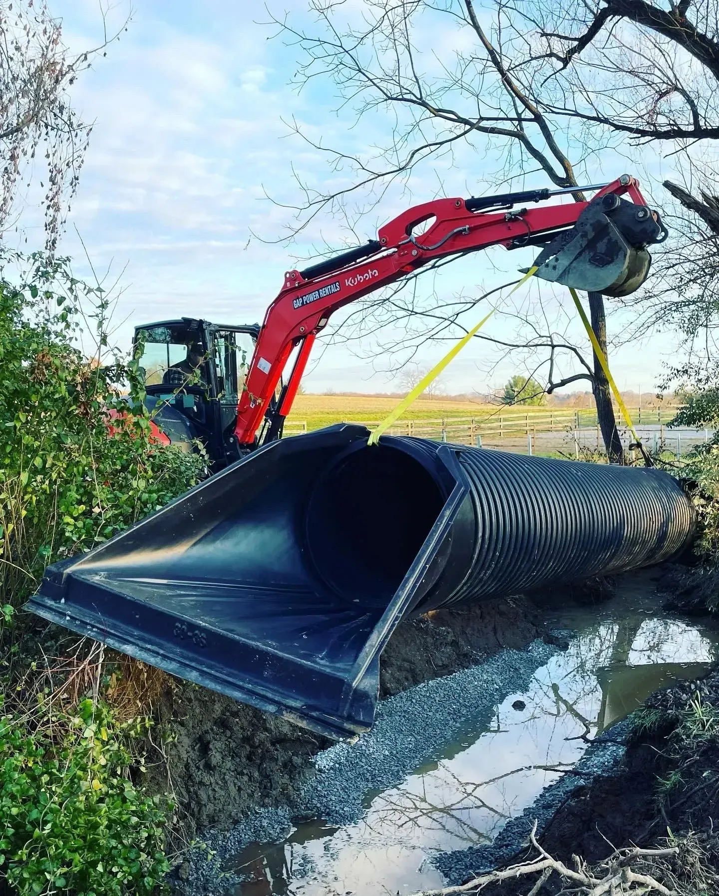A red excavator lifting a large, black, corrugated culvert pipe into a muddy drainage ditch surrounded by trees.