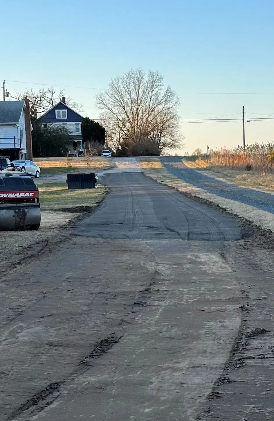 A road under construction, with a dark, newly paved section leading toward a house and a large tree at sunset.