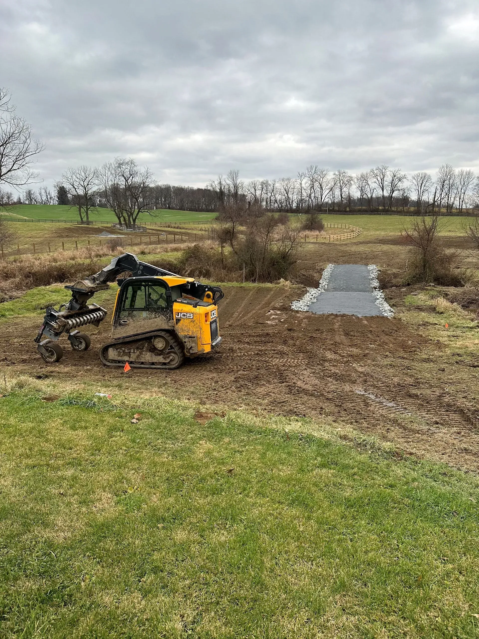 A yellow tracked skid steer loader works on muddy ground near a gray stone crossing in a field under a cloudy sky.