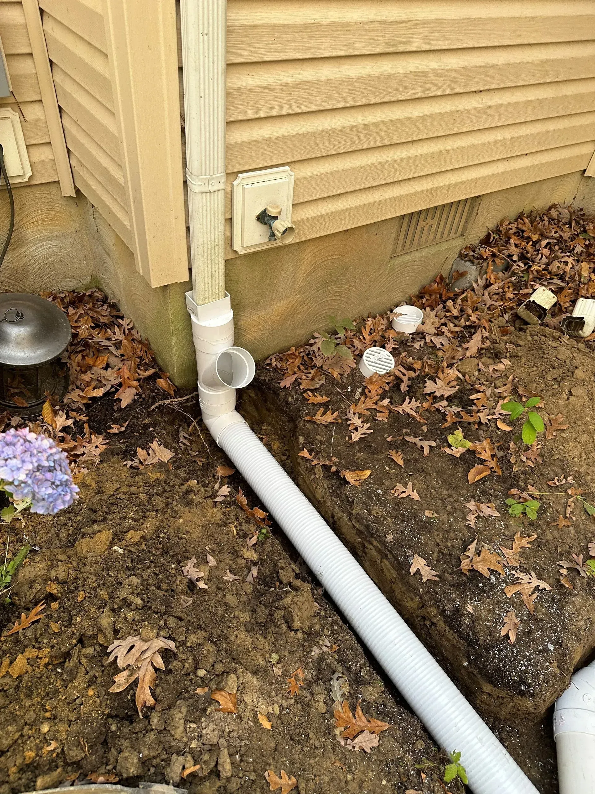 A white downspout extension connected to a PVC pipe running across a dirt garden bed next to a house foundation.