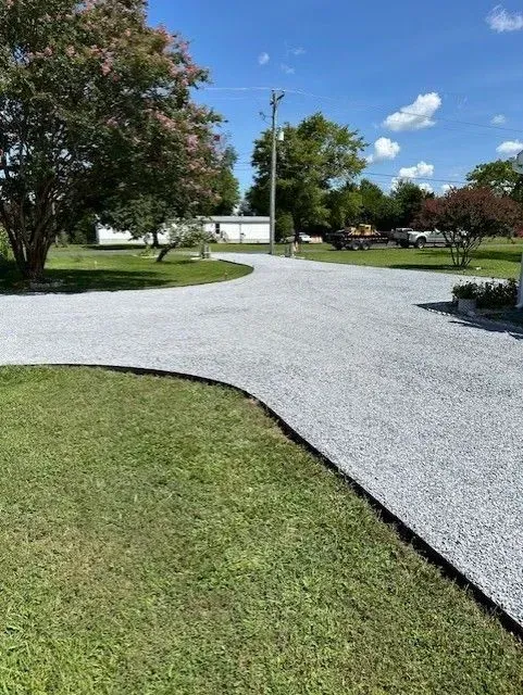 A gravel driveway curves through a sunny lawn toward a distant white house under a bright blue sky.