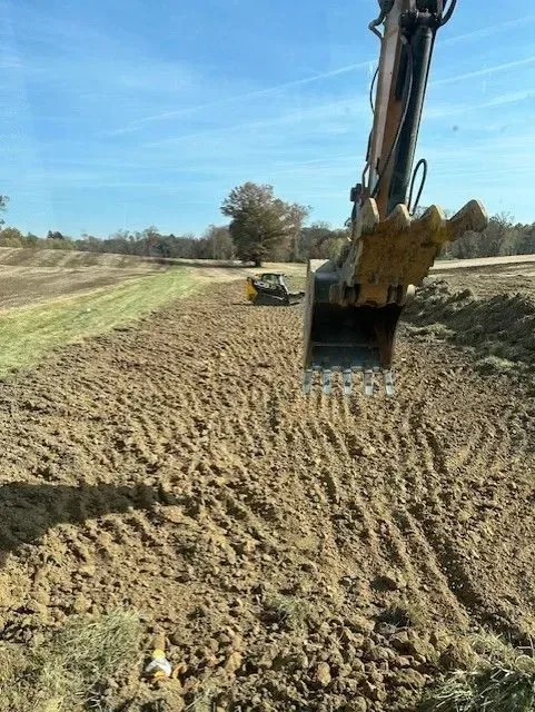 An excavator bucket is suspended in the foreground, overlooking a field with another piece of machinery in the distance.